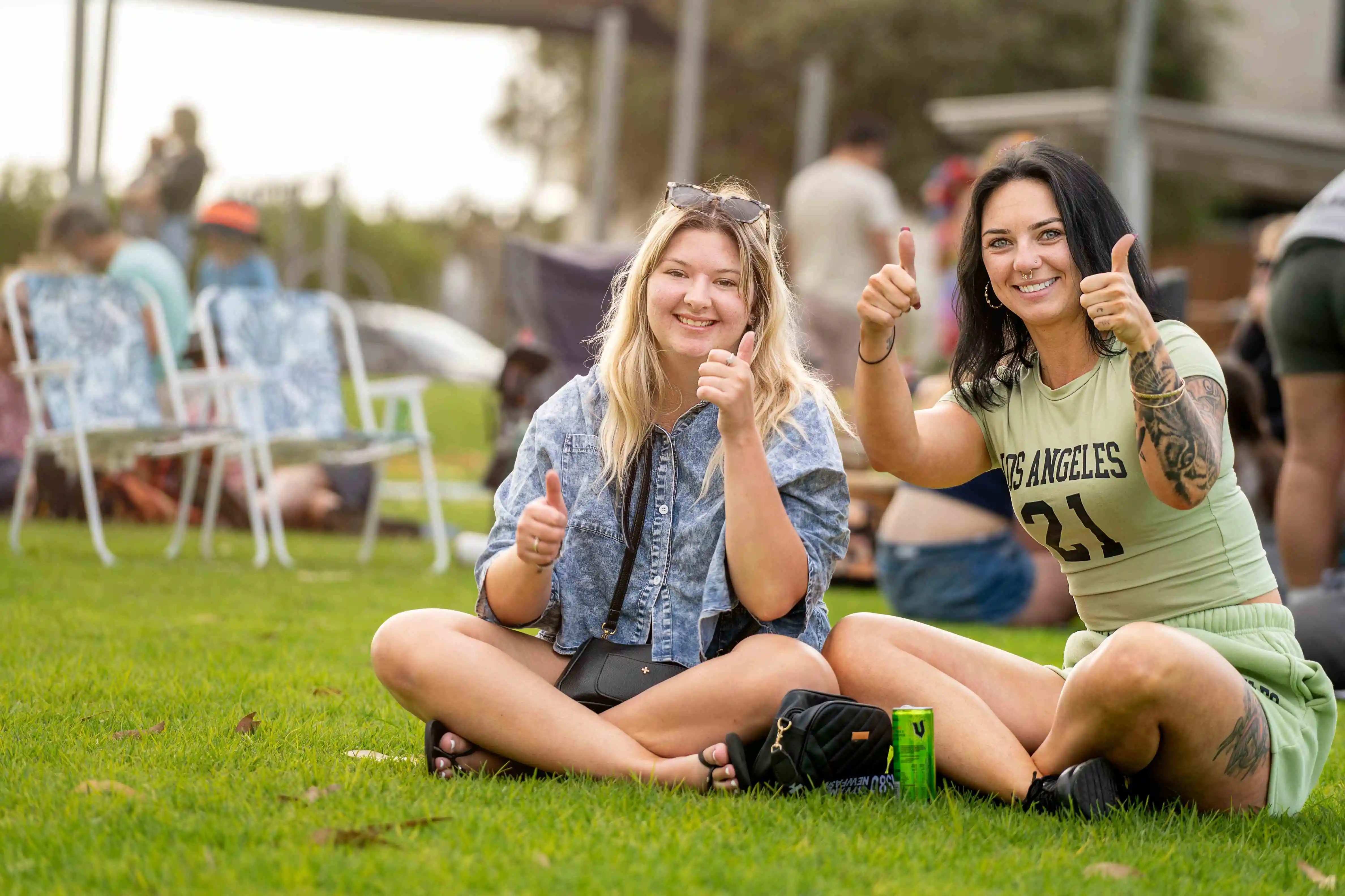 Two people sitting on grass at an outdoor event, both giving a thumbs‑up gesture. 