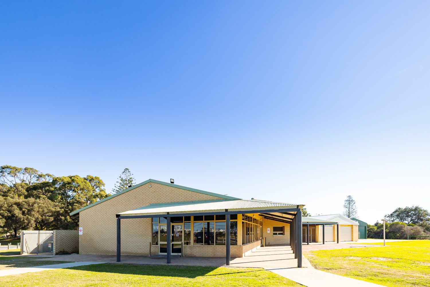 Halls Head Community Centre exterior with a single-storey brick building, covered entry walkway, large windows and surrounding grassed area under a clear blue sky.